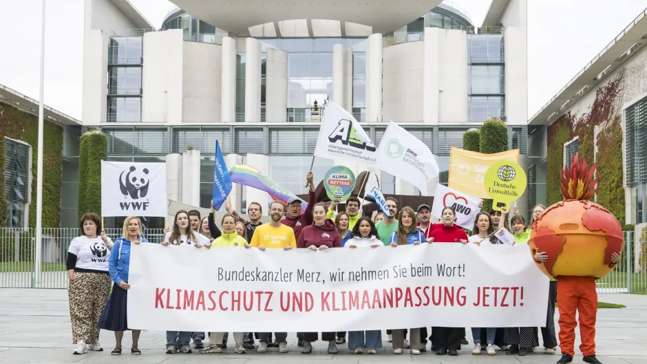 zwei Dutzend Umweltaktive vor dem Bundeskanzleramt in Berlin mit einem Banner, Aufschrift "Bundeskanzler Merz, wir nehmen Sie beim Wort! KLIMASCHUTZ UND KLIMAANPASSUNG JETZT!"; rechts im Bild, eine als brennende Erde verkleidete Person